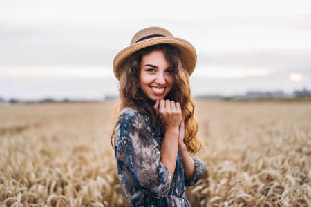 Closeup portrait of a beautiful young woman with curly hair. Woman in dress and hat standing in wheat field.の写真素材