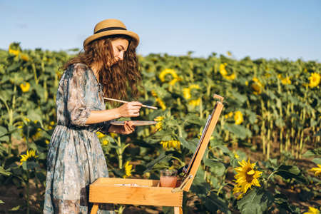 A young woman with curly hair and wearing a hat is painting in nature. A woman stands in a sunflower field on a beautiful day.の写真素材