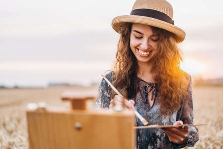 Beautiful girl artist paints in a wheat field. Woman smiles, holds a brush and palette in hands, in front of her stands an easel. Behind her is orange sunlight.の写真素材