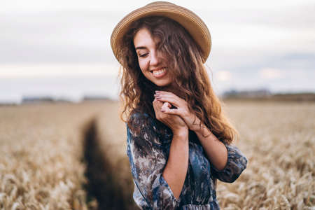Closeup portrait of a beautiful young woman with curly hair. Woman in dress and hat standing in wheat field.の写真素材