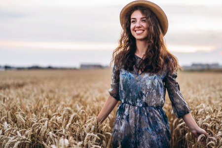 Closeup portrait of a beautiful young woman with curly hair. Woman in dress and hat standing in wheat field.の写真素材