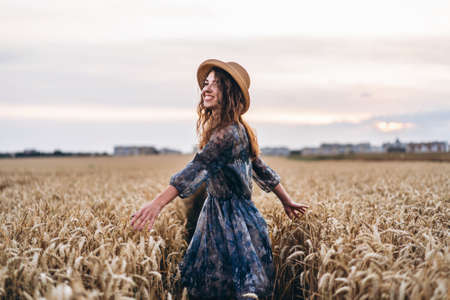 Closeup portrait of a beautiful young woman with curly hair. Woman in dress and hat standing in wheat field.の写真素材
