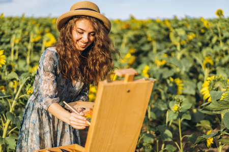 A young woman with curly hair and wearing a hat is painting in nature. A woman stands in a sunflower field on a beautiful day.の写真素材