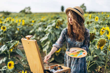 A young woman with curly hair and wearing a hat is painting in nature. A woman stands in a sunflower field on a beautiful day.の写真素材
