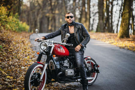 Bearded brutal man in sunglasses and leather jacket sitting on a motorcycle on the road in the forest with blured colorful background.の写真素材