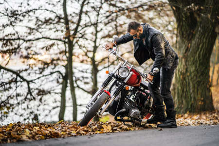 Bearded brutal man in sunglasses and leather jacket sitting on a motorcycle on the road in the forest with blured colorful background.の写真素材