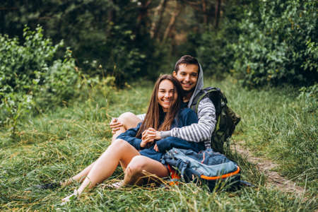Woman with long hair and man sitting on the grass in the forest with backpacks. Have fun in nature, hugging and kissing.の写真素材