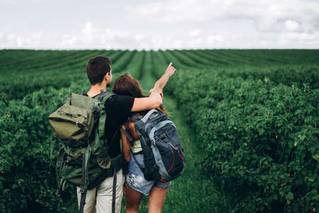 Back view of young couple with backpacks on currant plantations. Woman with long hair points her hand to the horizon.の写真素材