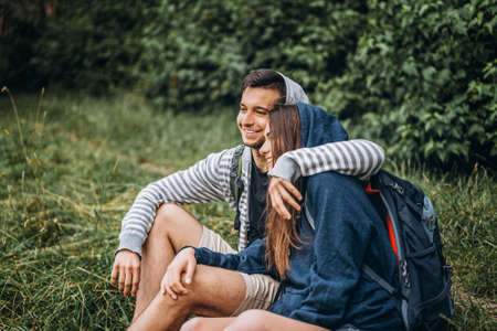 Woman with long hair and man sitting on the grass in the forest with backpacks. Have fun in nature, hugging and kissing.の写真素材