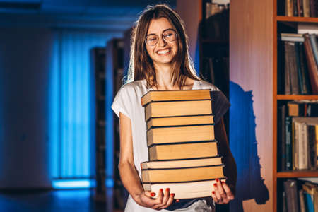Young girl student with glasses in library smiling and carries stack of books. Exam preparation.の写真素材