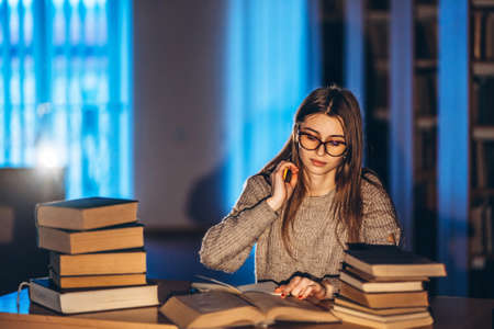 Young student in glasses preparing for the exam. Girl in the evening sits at a table in the library with a pile of books.の写真素材