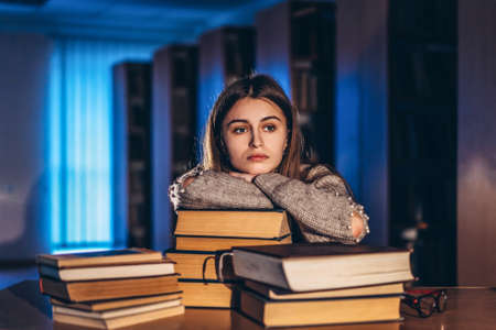 Tired student girl in the evening sitting leaning on his arm in the library at the desk with books. Teaching and preparation for exams.の写真素材