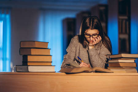 Young student in glasses preparing for the exam. Girl in the evening sits at a table in the library with a pile of books.の写真素材