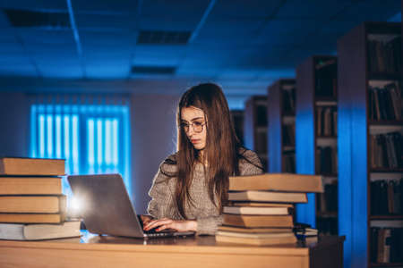 Young student woman in the evening sits at a table in the library with a pile of books and works on a laptop. Preparing for the exam.の写真素材