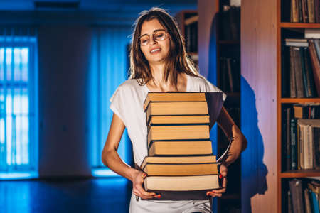 Young girl student with glasses in library smiling and carries stack of books. Exam preparation.の写真素材