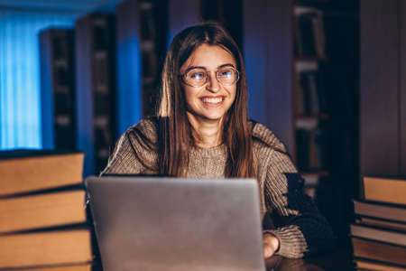 Young smiling student woman in the evening sits at a table in the library with a pile of books and works on a laptop. Preparing for the exam.の写真素材