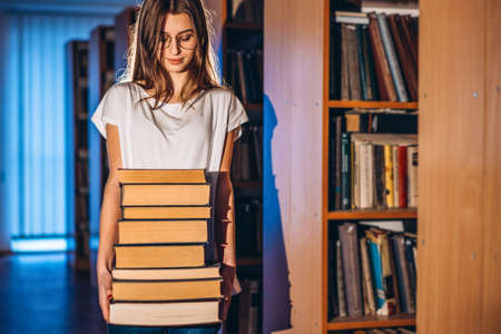 Young girl student with glasses in library smiling and carries stack of books. Exam preparation.の写真素材