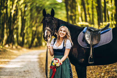 Pretty blonde in traditional dress  walking with big black horse in the forest. Animal and people frienship concept.の写真素材