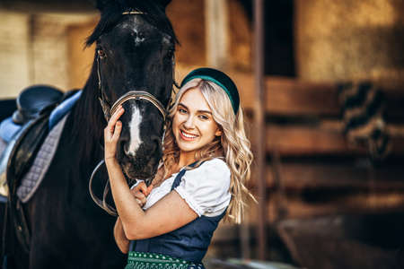 Pretty blonde in traditional dress  take care of big black horse at the farm. Animal and people frienship concept.の写真素材