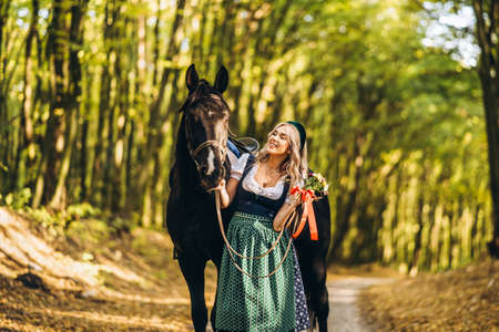 Pretty blonde in traditional dress  walking with big black horse in the forest. Animal and people frienship concept.の写真素材