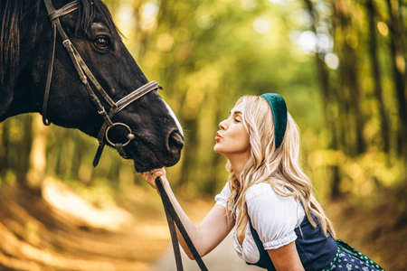 Pretty blonde in traditional dress  walking with big black horse in the forest. Animal and people frienship concept.の写真素材