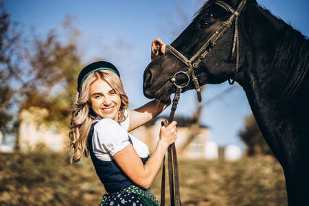 Pretty blonde in traditional dress  walking with big black horse outdoors. Animal and people frienship concept.の写真素材