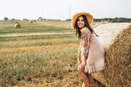 Beautiful young girl with long hair in sunnglasses and straw hat posing on a wheat field near hay bales. Happy brunette in summer dress.の写真素材
