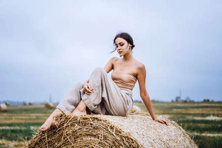 Brunette in linen pants and bare shoulders sitting on a hay bales in warm autumn day. Woman looking at camera. Behind her is a wheat field.の写真素材