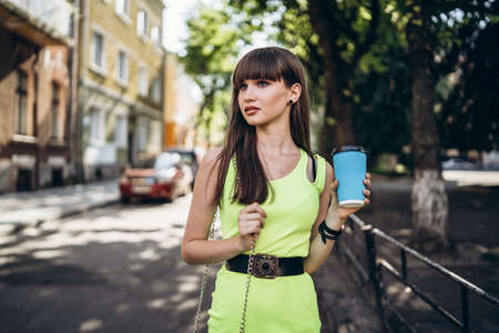 Pretty long hair brunette girl in green light dress with blue paper cup of beverage walking outdoor on the street in the cityの写真素材