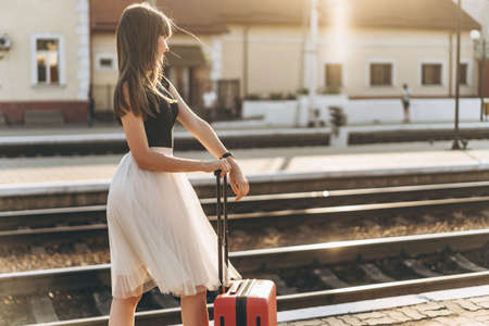 Female brunette traveler with red suitcase in white skirt waiting for a train and walking on raiway stationの写真素材
