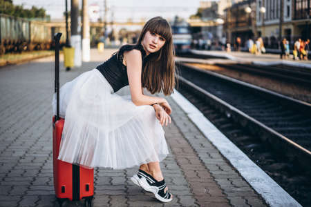 Female brunette traveler with red suitcase in white skirt waiting for a train on raiway stationの写真素材