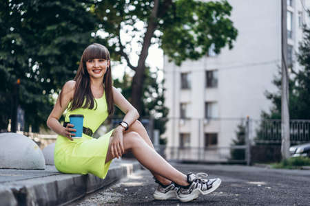 Pretty long hair brunette girl in green light dress with blue paper cup of beverage sitting outdoor on the street in the cityの写真素材