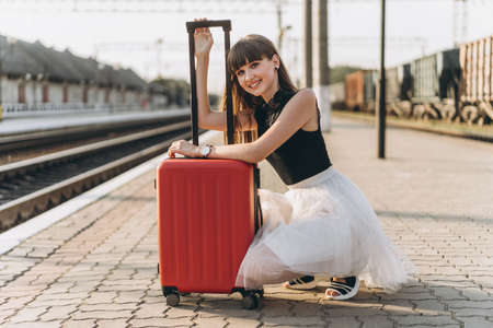 Female brunette traveler with red suitcase in white skirt waiting for a train on raiway stationの写真素材