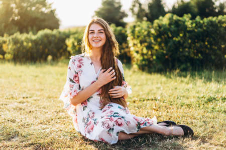 Full length portrait of beautiful woman with long curly hair on currant field background. Girl in a light dress sits on the grass in sunny day.の写真素材