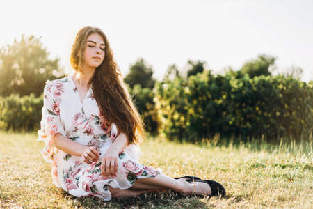 Full length portrait of beautiful woman with long curly hair on currant field background. Girl in a light dress sits on the grass in sunny day.の写真素材