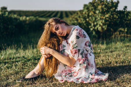 Full length portrait of beautiful woman with long curly hair on currant field background. Girl in a light dress sits on the grass in sunny day.の写真素材
