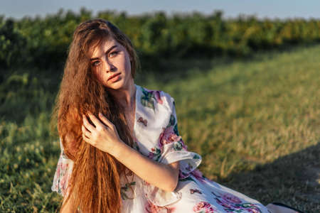 Young girl with long curly hair, brunette, face in freckles, with makeup and green eyes, in light white dress posing for camera, background blurred, natureの写真素材
