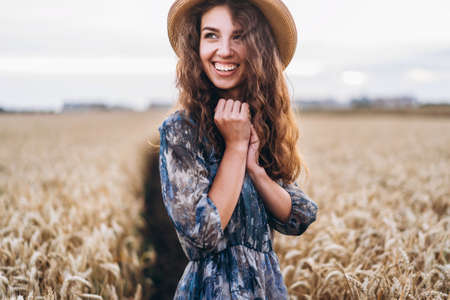 Closeup portrait of a beautiful young woman with curly hair. Woman in dress and hat standing in wheat field.の写真素材