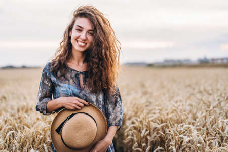 Closeup portrait of a beautiful young woman with curly hair. Woman in dress and hat standing in wheat field.の写真素材