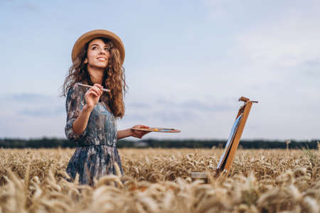 Portrait of smiling female artist with curly hair in hat. Girl draws a picture of a landscape in a wheat field. Copy space.の写真素材
