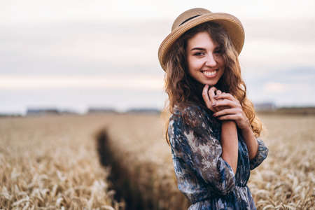 Closeup portrait of a beautiful young woman with curly hair. Woman in dress and hat standing in wheat field.の写真素材