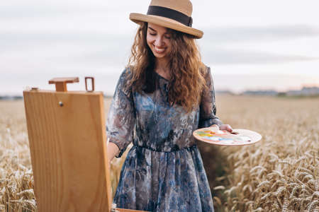Portrait of smiling female artist with curly hair in hat. Girl draws a picture of a landscape in a wheat field.の写真素材