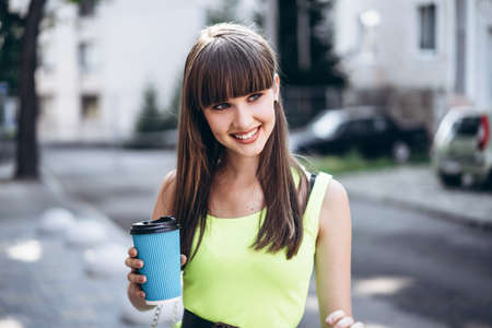 Pretty long hair brunette girl in green light dress with blue paper cup of beverage walking outdoor on the street in the cityの写真素材
