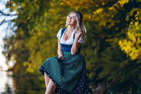 Oktoberfest, St. Patrick day, intenational beer day. Happy pretty blond girl in dirndl, traditional beer festival dress, sitting outdoors with blured colorful trees behindの写真素材