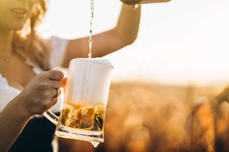 Close-up of the girls hands in dirndl, traditional festival dress, pouring a full glass of beer with huge foam, outdoors with blurred background. Oktoberfest, St. Patrickâs day, international beer day conceptの写真素材
