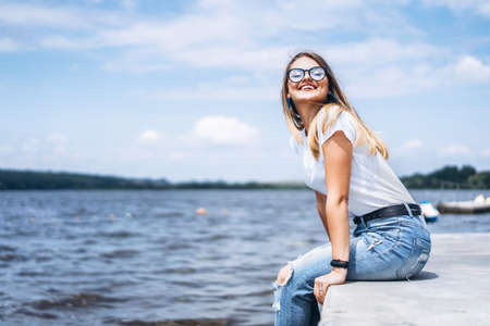 Young woman with long hair in stylish glasses posing on the concrete shore near the lake. Girl dressed in jeans and t-shirt smiling and looking at the camera.の写真素材