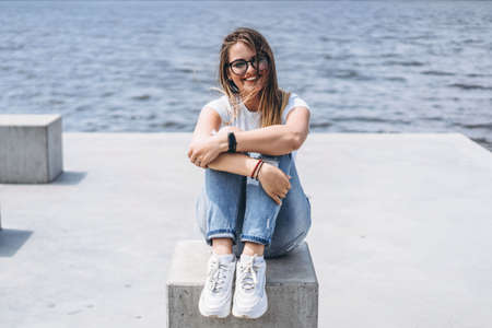 Young woman with long hair in stylish glasses posing on the concrete shore near the lake. Girl dressed in jeans and t-shirt smiling and looking away.の写真素材