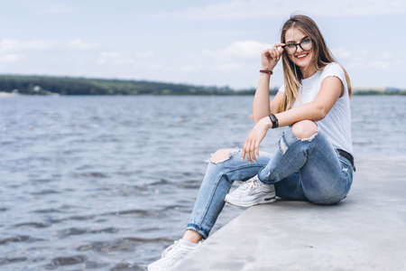 Young woman with long hair in stylish glasses posing on the concrete shore near the lake. Girl dressed in jeans and t-shirt smiling and looking at the camera.の写真素材
