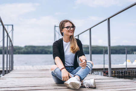 Young woman with long hair in stylish glasses posing on a wooden pier near the lake. Girl dressed in jeans and t-shirt smiling and looking awayの写真素材