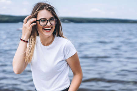 Portrait of a beautiful young woman in stylish glasses. Girl in white tshirt posing on the background of the lake landscape.の写真素材